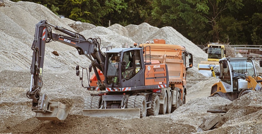 Baustellengeräte auf einer Baustelle. Im Vordergrund ein Bagger und ein Kipplaster dahinter auf einem Kiesberg. 