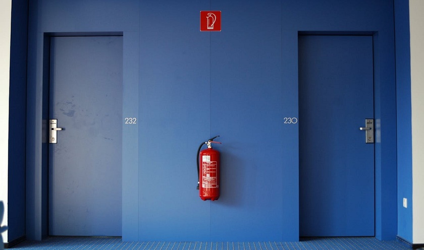 Zwei blaue Türen in einer blauen Wand. Dazwischen ein Feuerlöscher, der an der Wand befestigt ist und das entsprechende Hinweiszeichen darüber. 