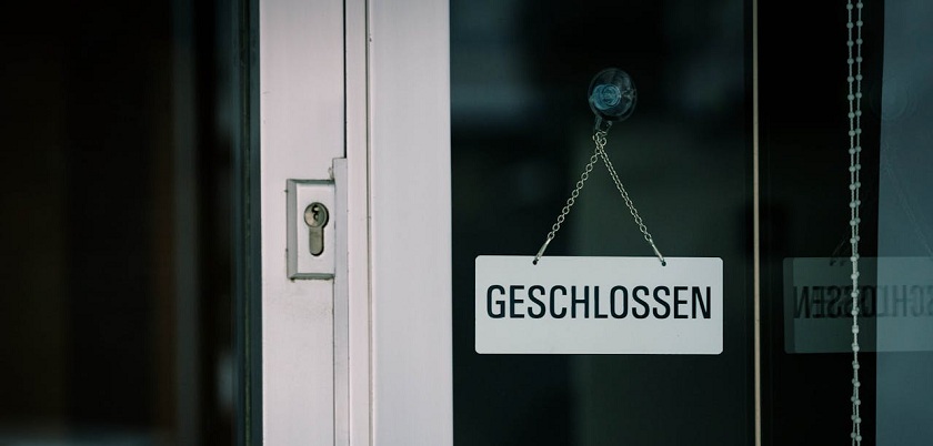 Teilausschnitt einer Eingangstür, in der ein weißes Schild mit der Aufschrift GESCHLOSSEN hängt 
